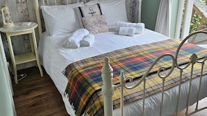 A neatly made bed with white linens, a plaid blanket, and decorative pillows. There is a small bedside table with a lamp, and a window with curtains. - Hooray Henrys Sheringham (Sheringham)