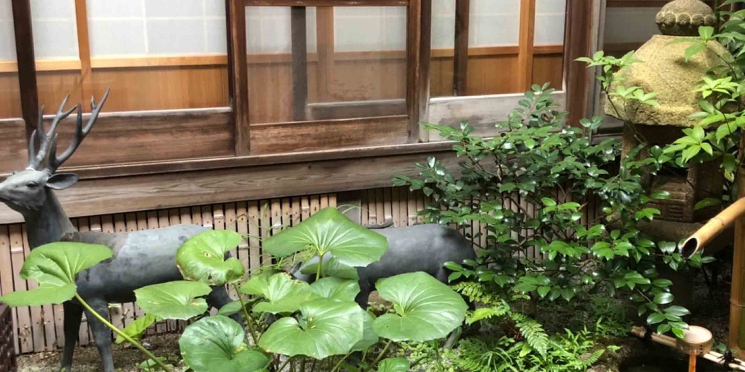 Un jardín con una estatua de ciervo, grandes plantas verdes y un farol tradicional japonés.