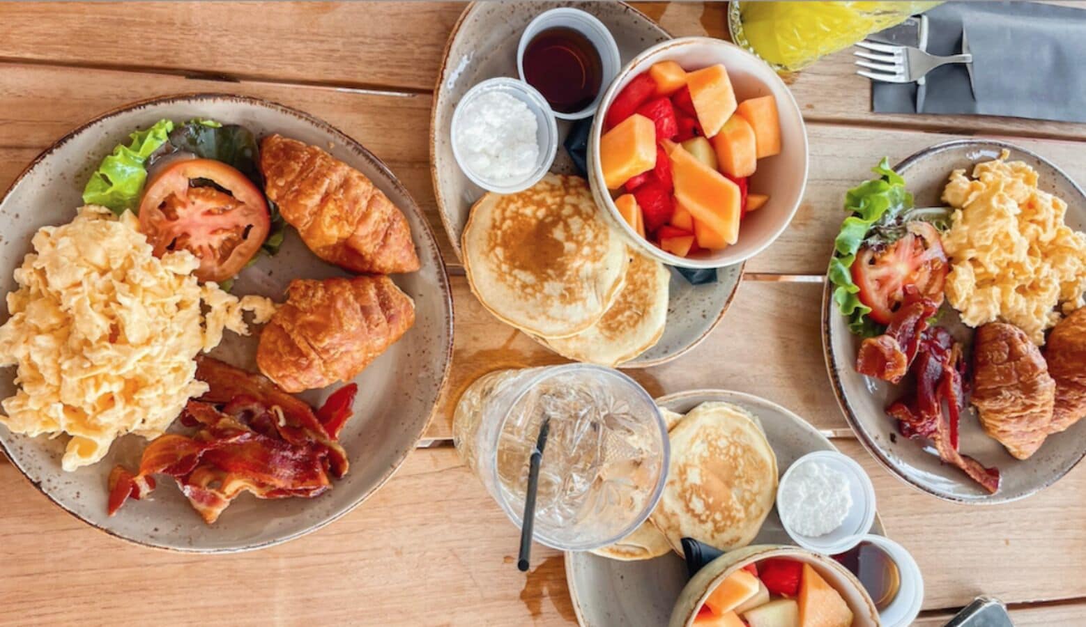 A breakfast spread with scrambled eggs, bacon, croissants, pancakes, and fruit on a wooden table.