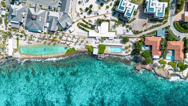 Luchtbeeld van een kustresort met heldere turquoise zee, een rotsachtige kustlijn en meerdere gebouwen, waaronder een hotel met zwembad en een restaurant.