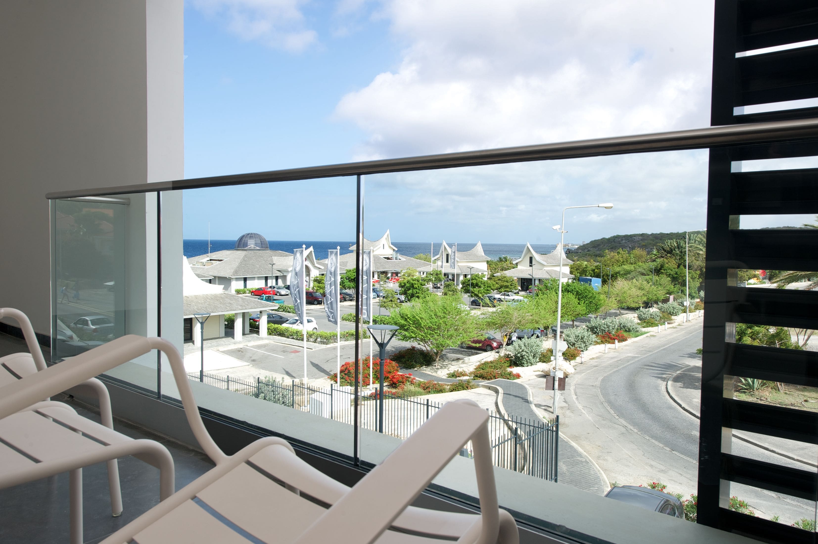 A balcony with a view of a coastal area, including a road, parked cars, and buildings with unique architecture.