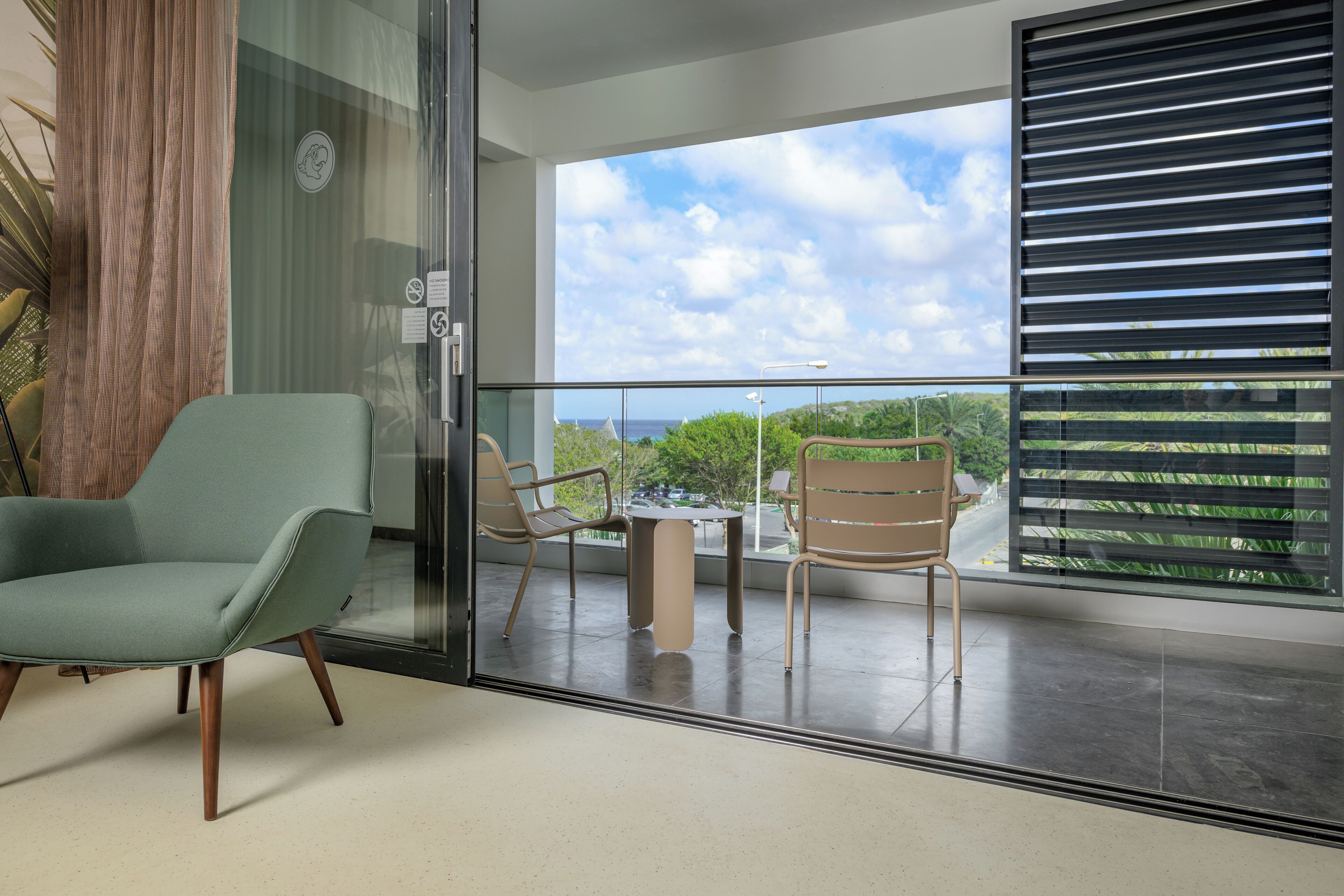A modern room with a sliding glass door leading to a balcony with a view of the sea. There is a green chair and a table with two chairs on the balcony.