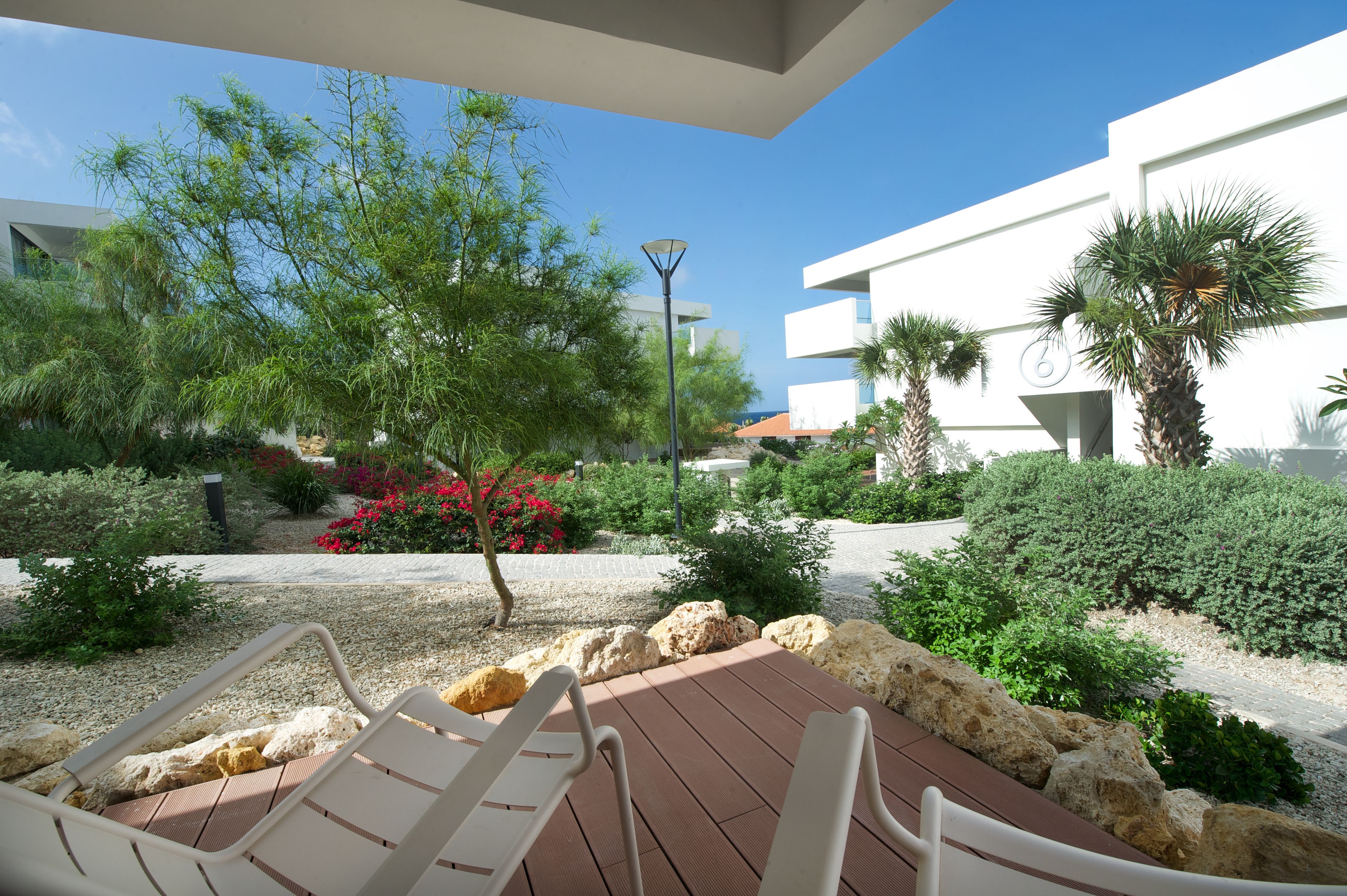 Un patio con sillones de descanso, un árbol pequeño y un jardín bien cuidado con diversas plantas y una decoración de rocas.