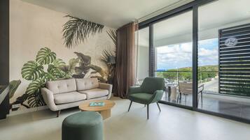 Una sala de estar moderna con sofá, sillón y mesa de centro, ventanas amplias con vista al exterior, y obras de arte con motivos de plantas tropicales en la pared.