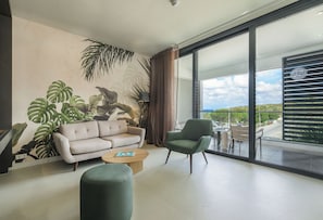 A modern living room with a sofa, armchair, and coffee table, large windows with a view of the outdoors, and tropical plant artwork on the wall.