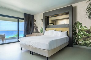 A modern hotel room with a large bed, a balcony with a view of the ocean, and tropical wall art.