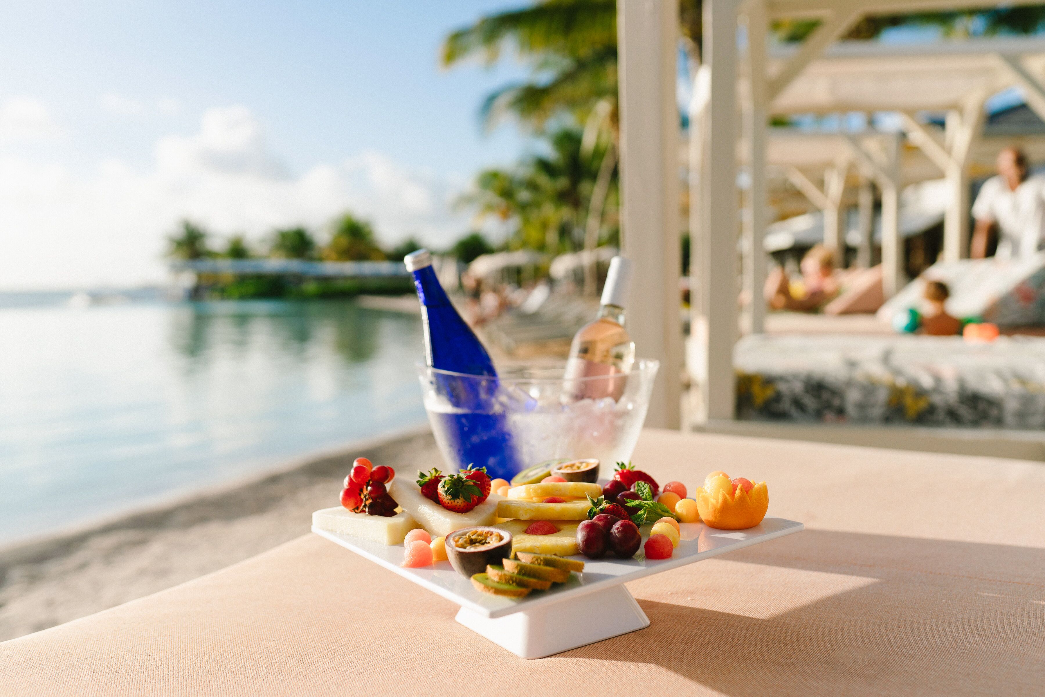 A table with a fruit platter and a bottle of water, overlooking a beach with palm trees and a pool.