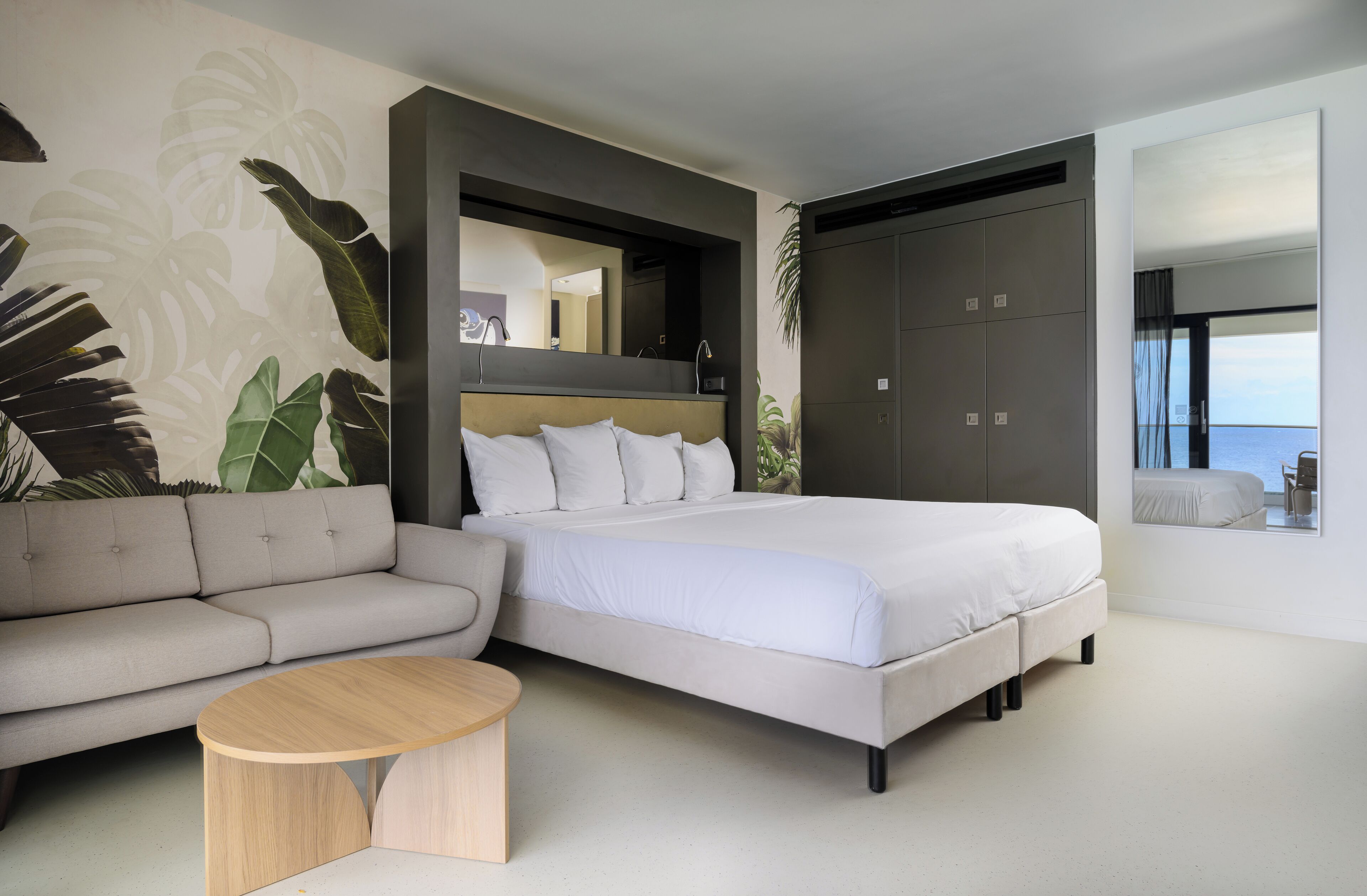 A modern bedroom with a large bed, a sofa, a round coffee table, and a view of the sea through a window.