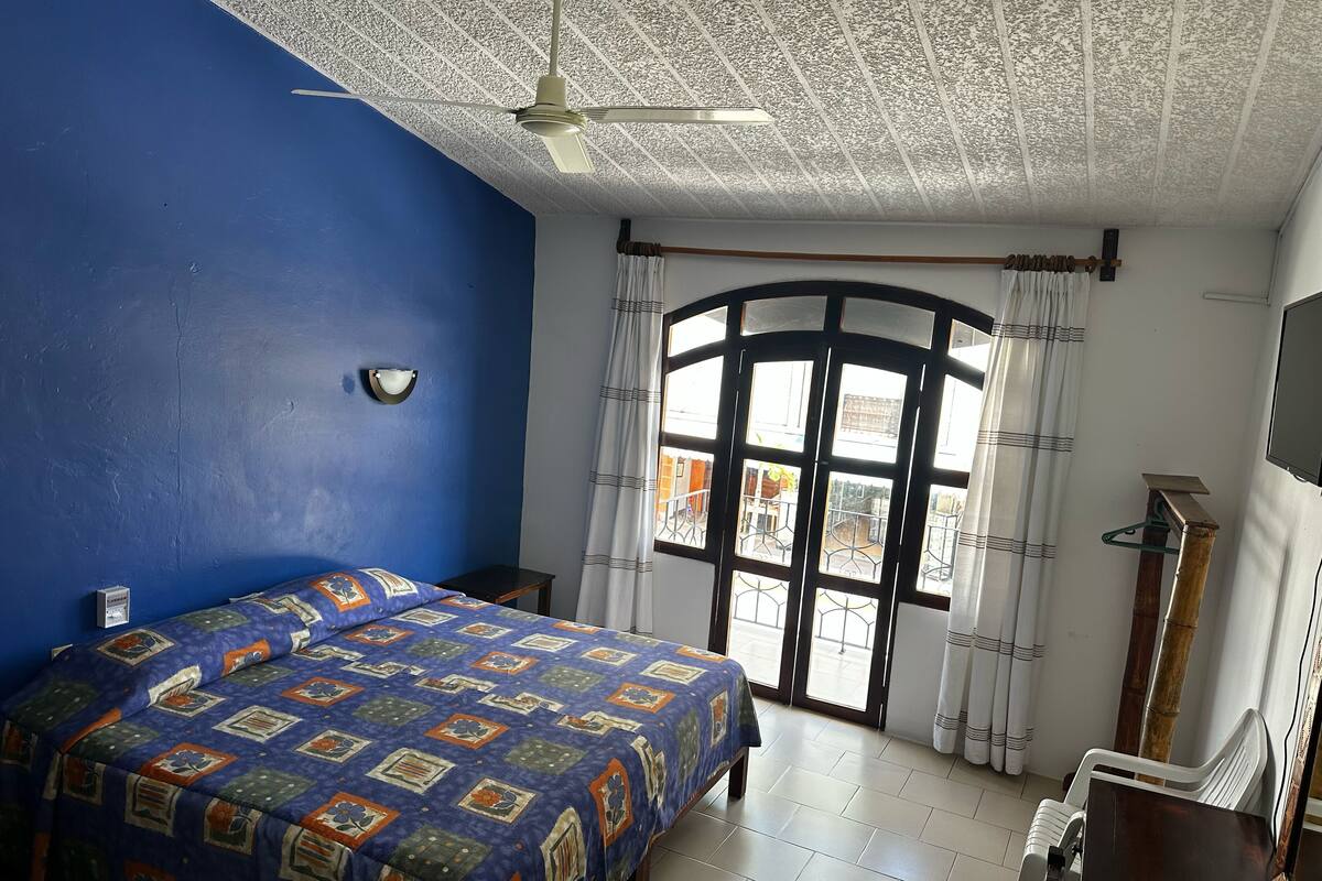 A bedroom with a large bed, a ceiling fan, a window with curtains, and a wooden nightstand.