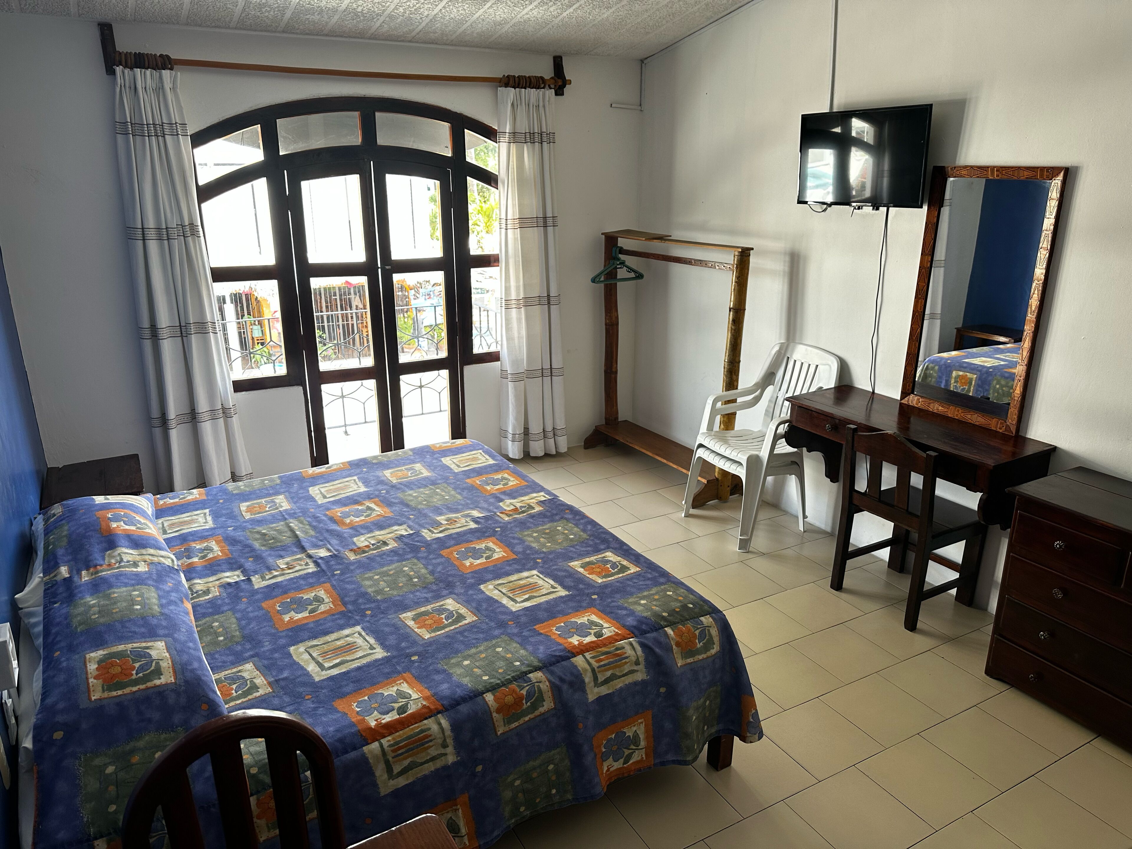 Une chambre à coucher comprenant un lit, un bureau, une chaise, une commode, un téléviseur et une fenêtre avec des rideaux.