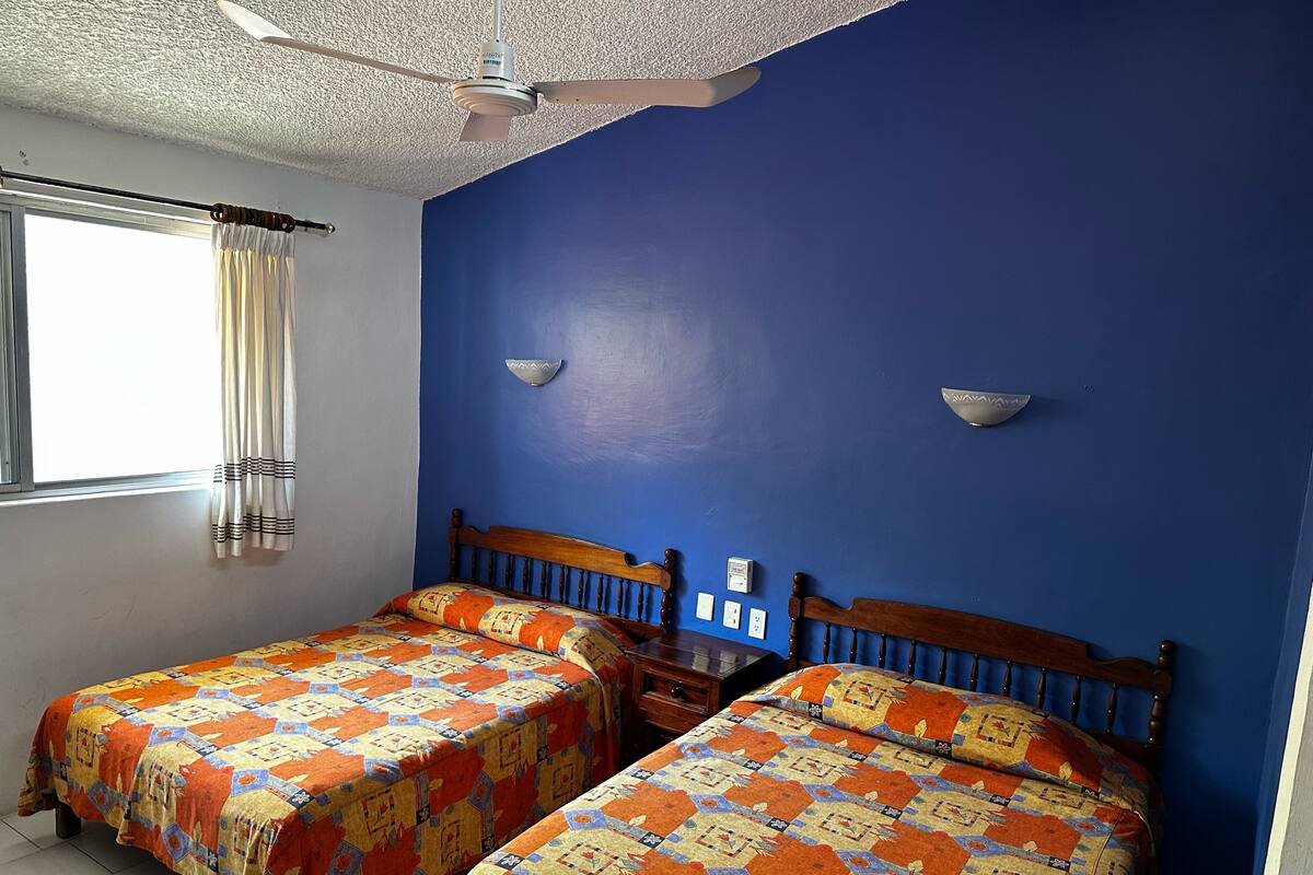 A room with two beds, a ceiling fan, a window with curtains, and wall-mounted lights.