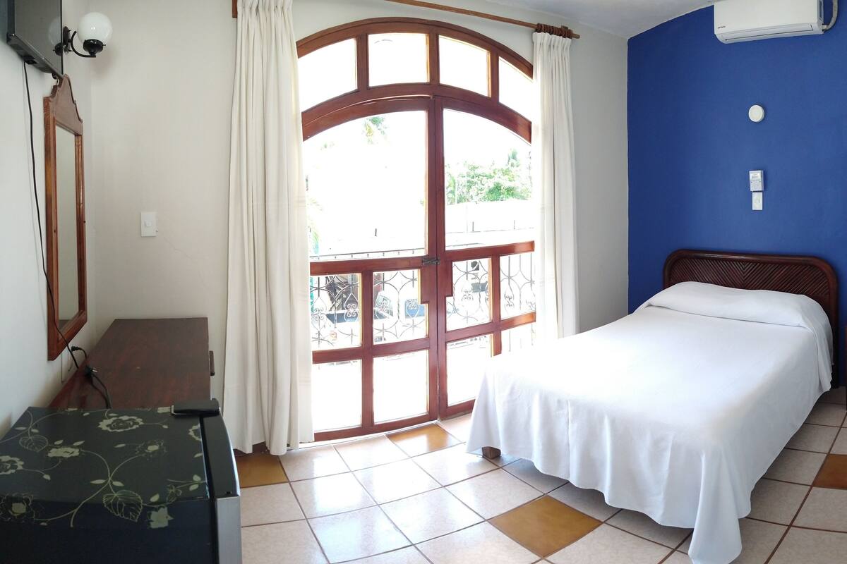 A bedroom with a bed, a desk, a chair, a window with curtains, and a wall-mounted air conditioner.
