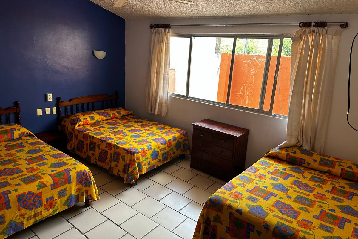 A room with two beds, a small wooden dresser, a ceiling fan, and a window with curtains.