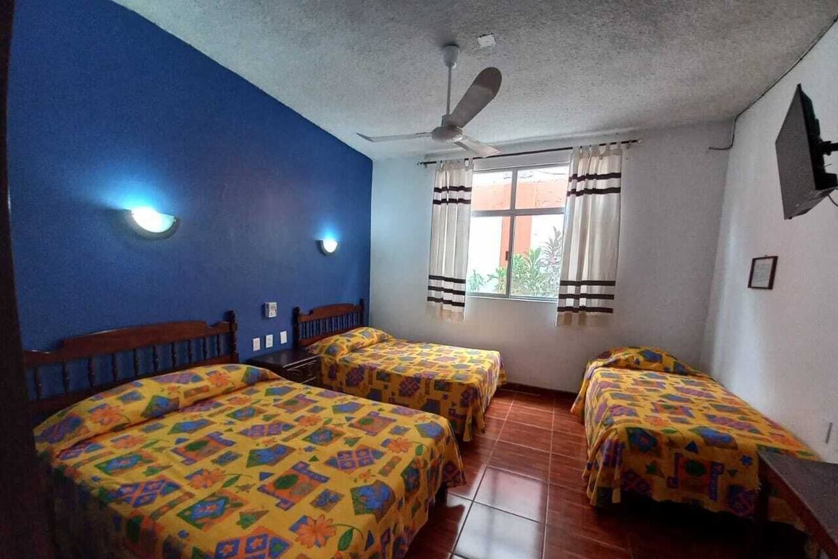 A room with four beds, a ceiling fan, a window with curtains, and a wall-mounted light.