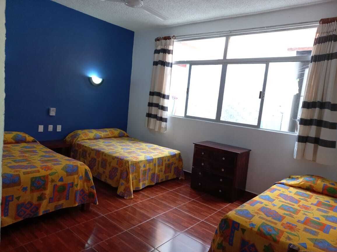 Una habitación con tres camas, un tocador, una ventana con cortinas y una lámpara fijada en la pared.