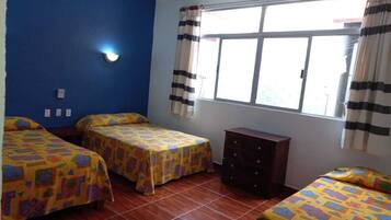 Una habitación con tres camas, un tocador, una ventana con cortinas y una lámpara fijada en la pared.