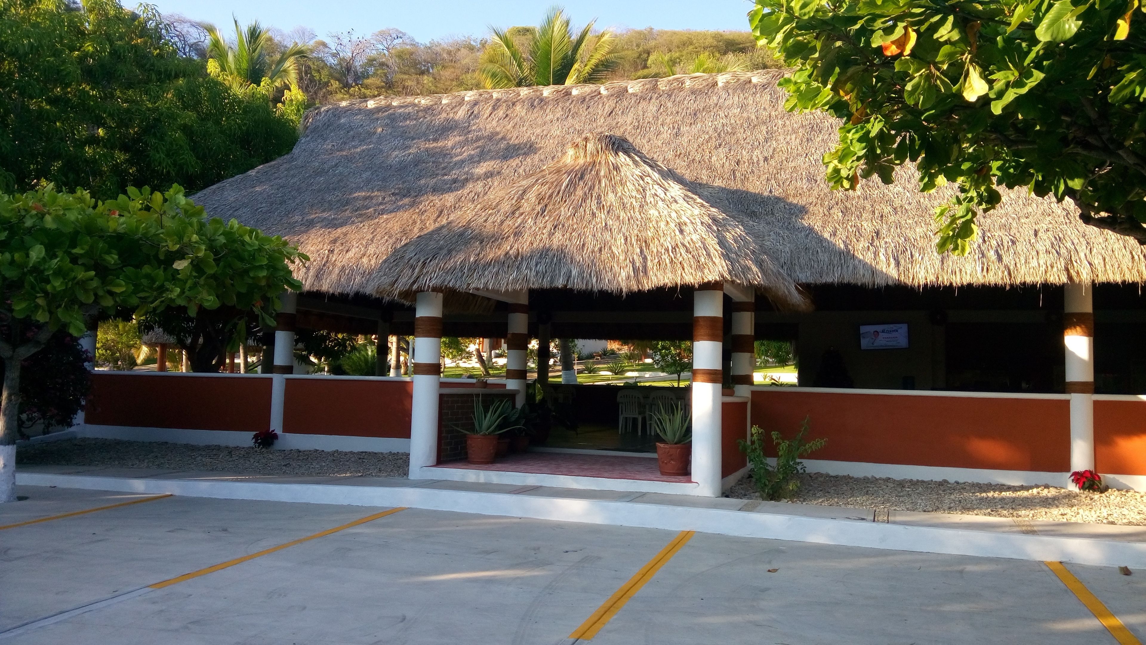 Un pabellón con techo de paja y zona cubierta para sentarse, rodeado de un estacionamiento y vegetación.