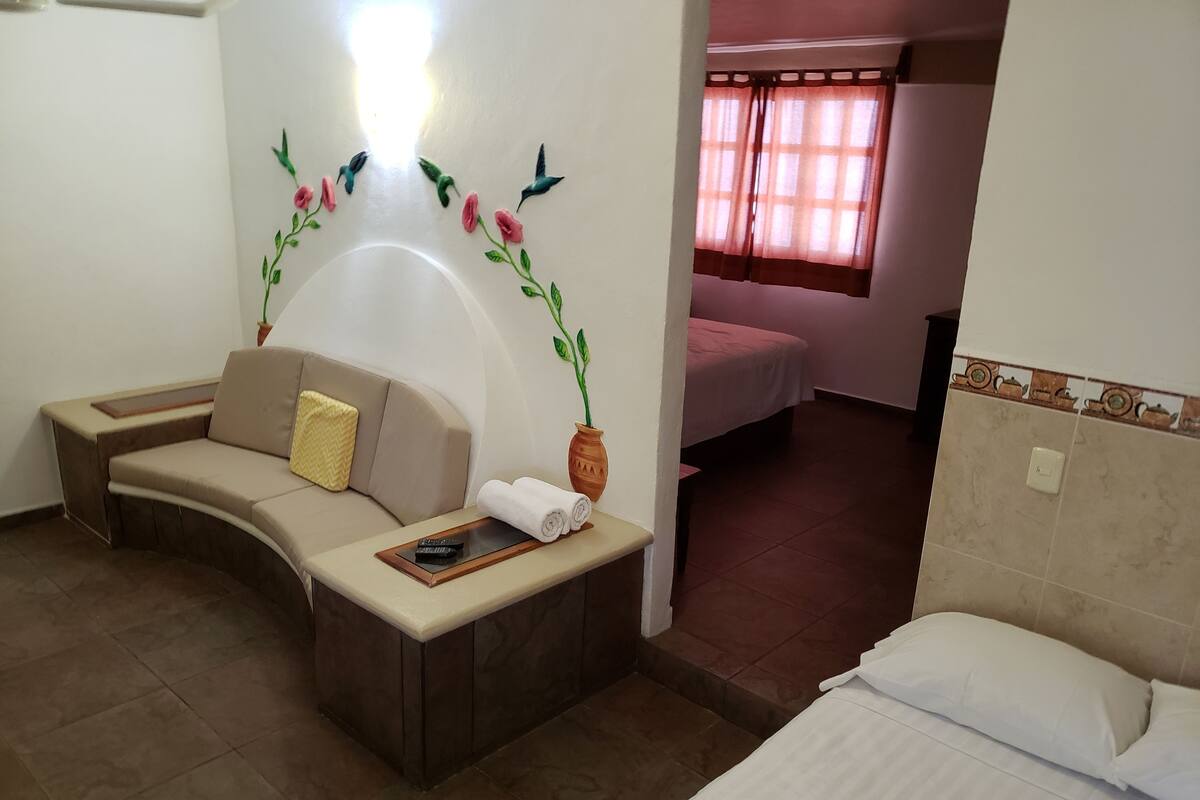 A hotel room with a sofa, a small table with a remote control, a potted plant, a bed with white bedding, and a window with curtains.
