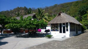 A small, white building with a thatched roof, surrounded by lush greenery and palm trees. - Hotel & Club Campestre Altos Paraiso (Villa de Tututepec de Melchor Ocampo)