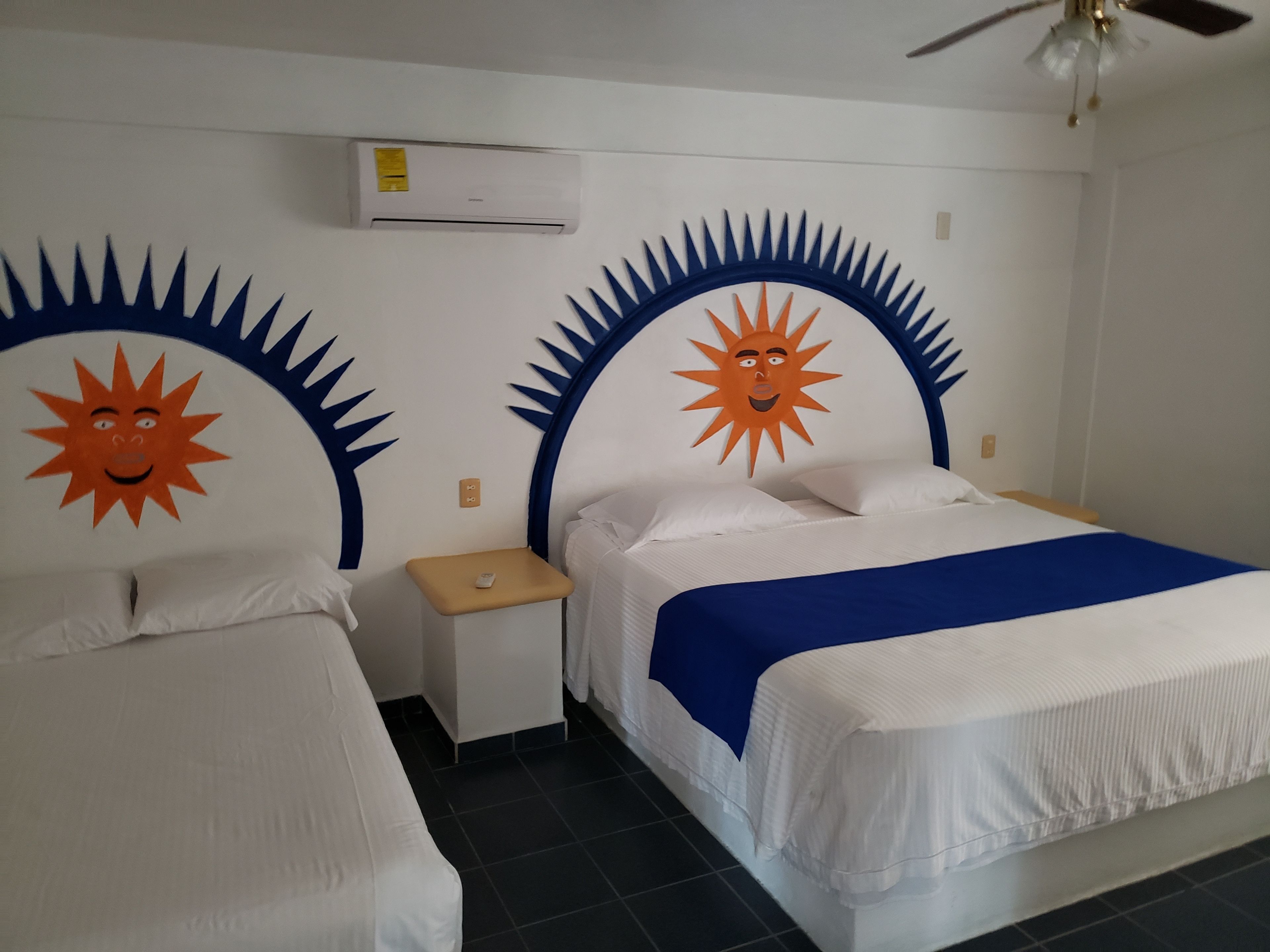 A hotel room with two beds, each with a sun design on the headboard, a small bedside table, an air conditioning unit above the beds, and a ceiling fan.