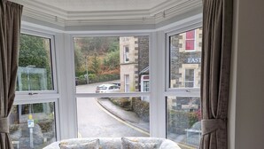 A corner room with a floral sofa, a bed, and large windows offering a view of the street and buildings outside. - Bowness Guest House (Windermere)