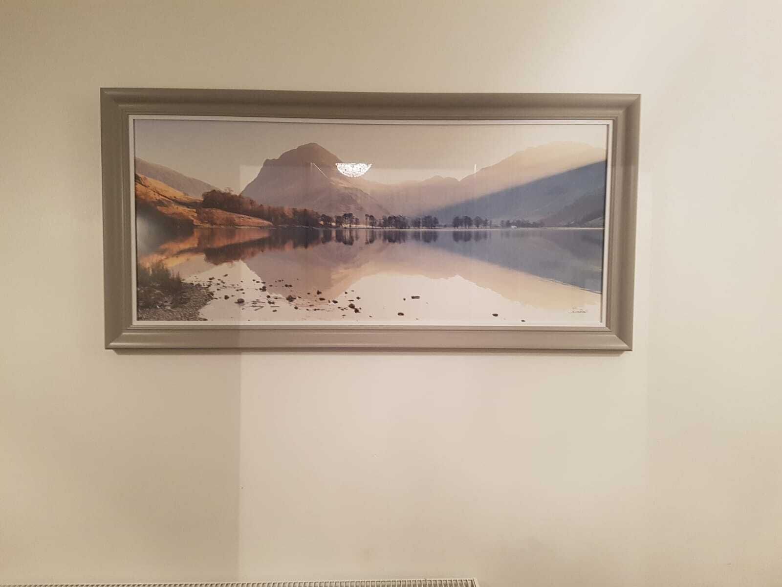A framed landscape painting of a calm lake with mountains and trees.
