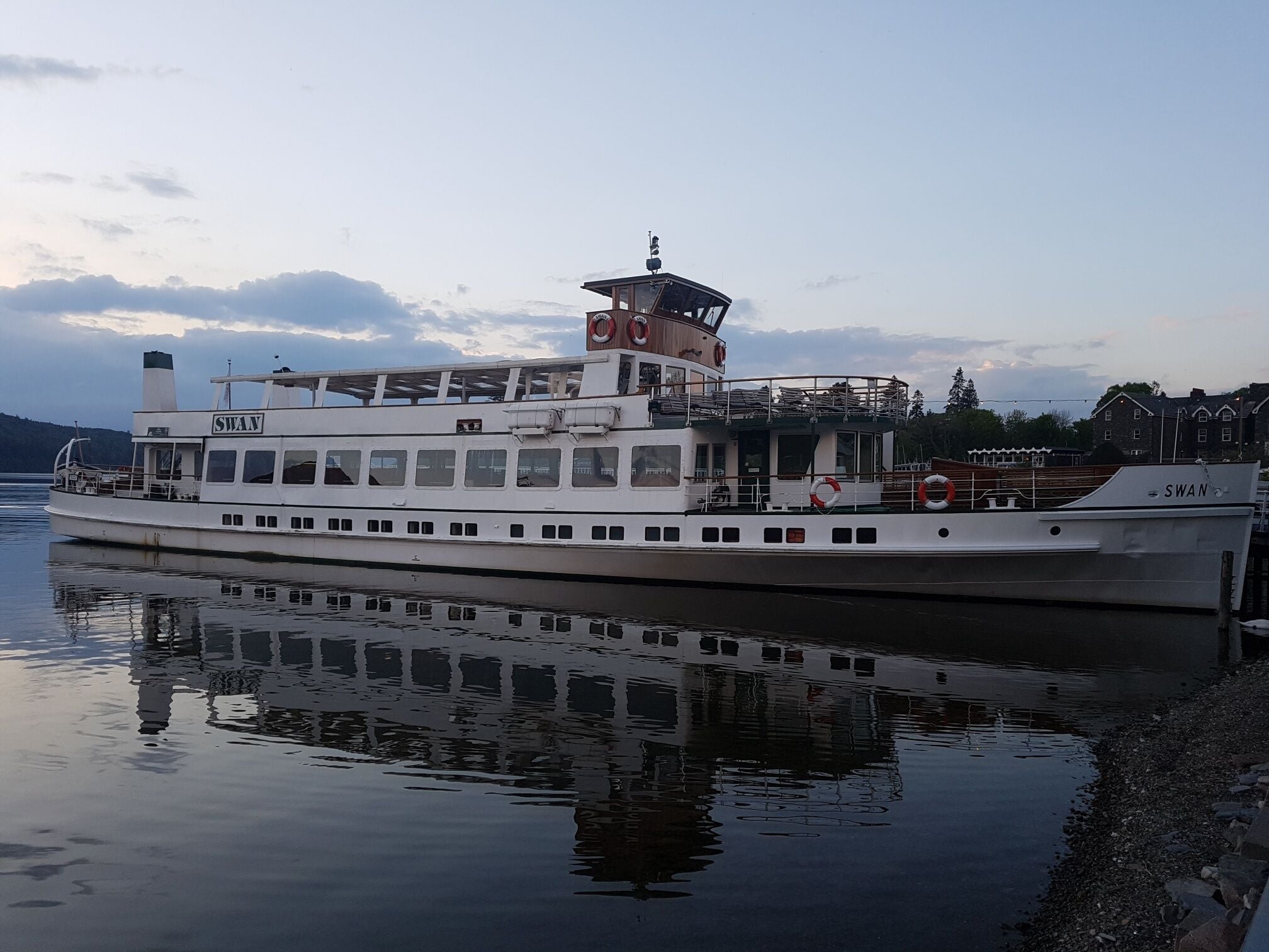 A white boat named 'SWAN' docked at a pier with a calm water surface reflecting the boat and the sky.