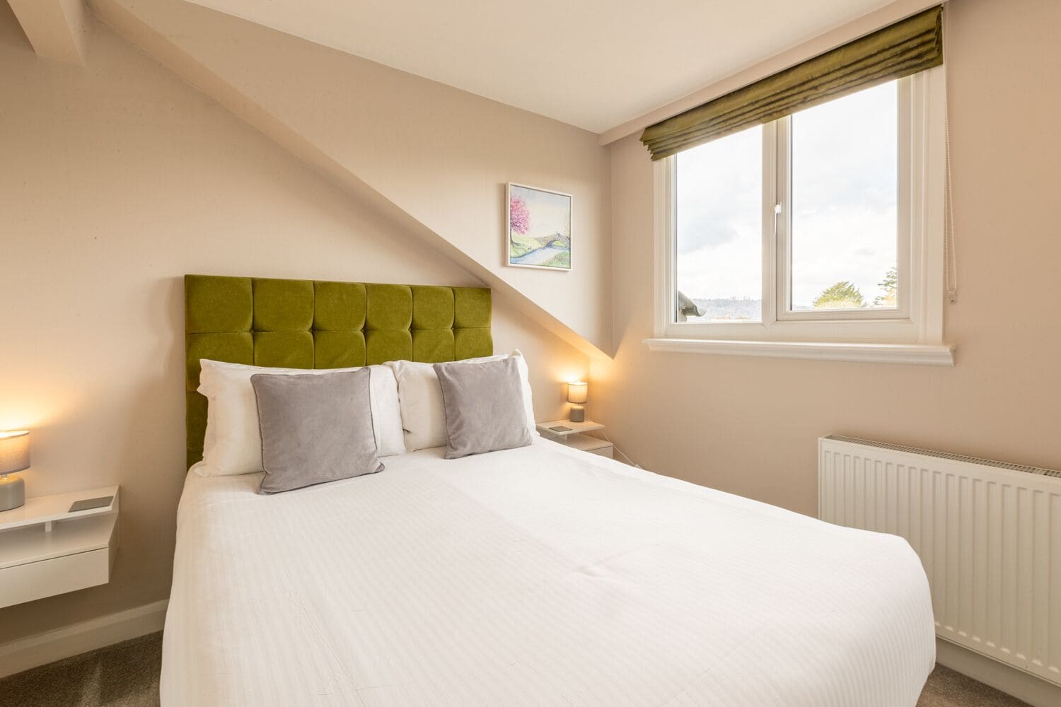 A bedroom with a green upholstered headboard, white bedding, and a window with a view of the outdoors.