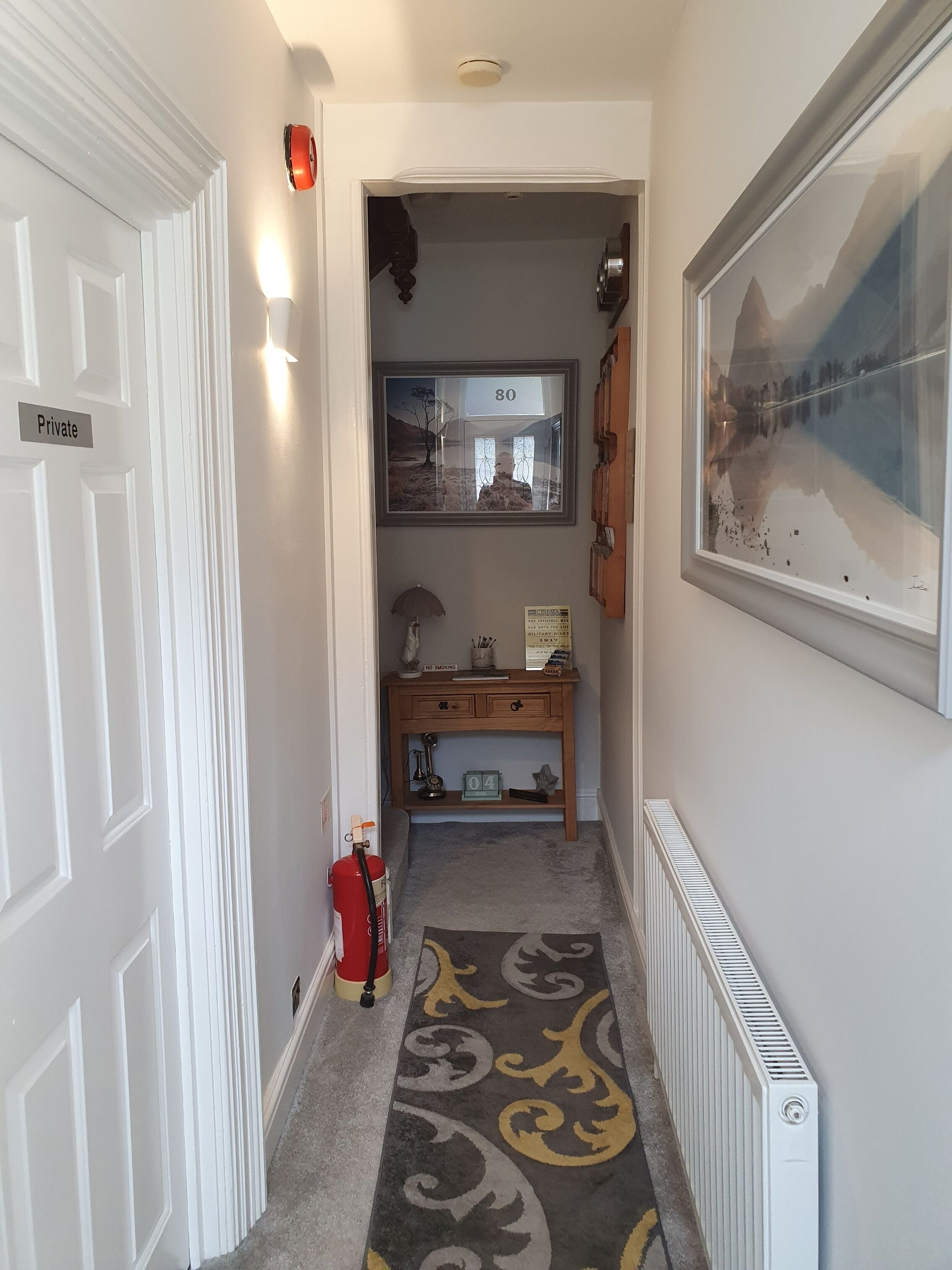 A hallway with a white door labeled "Private," a framed picture on the wall, a wooden desk with a "No Smoking" sign, and a radiator on the floor.