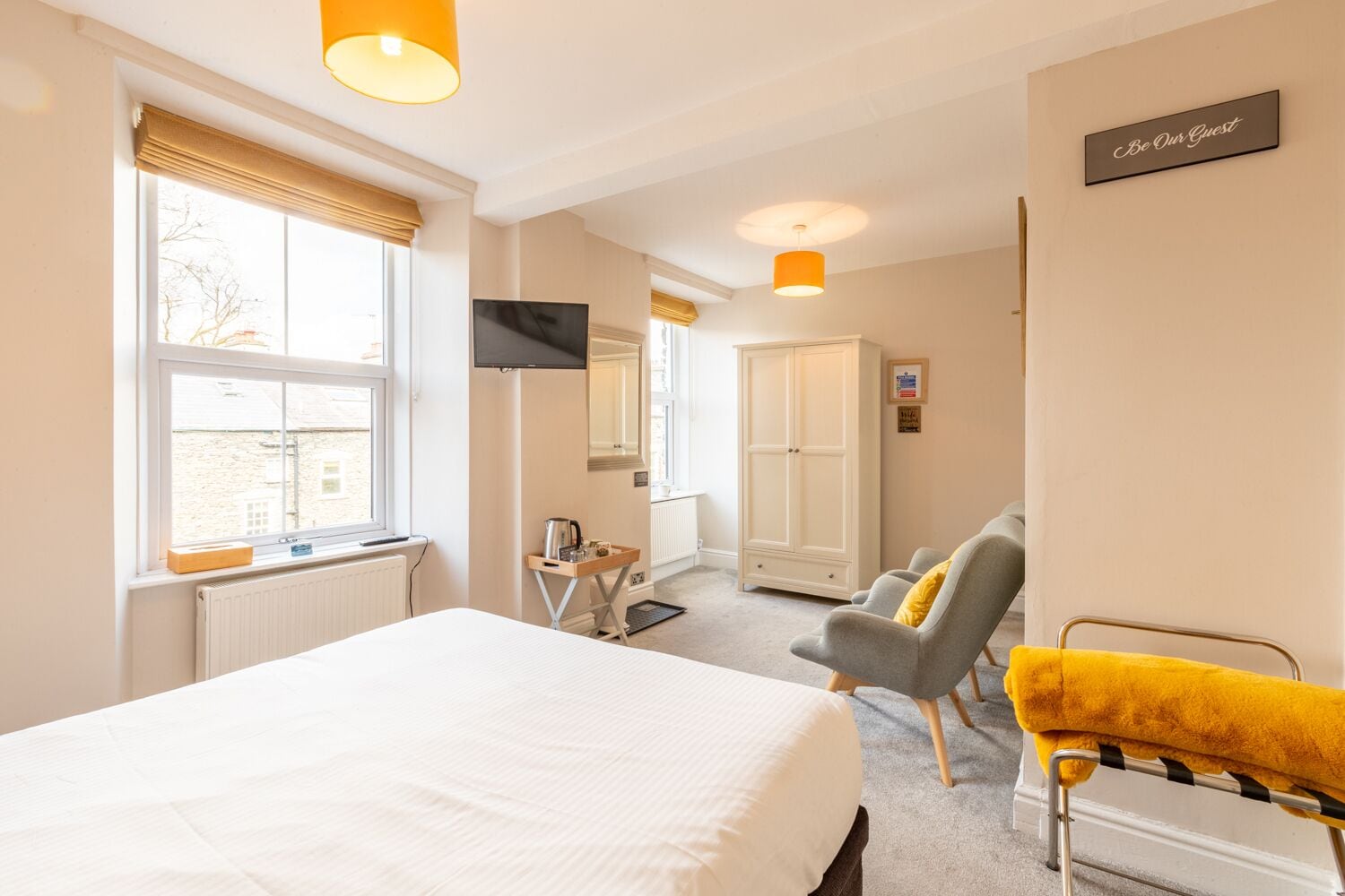A hotel room with a large bed, a TV, a wardrobe, a chair, and a sign that says "Be Our Guest".