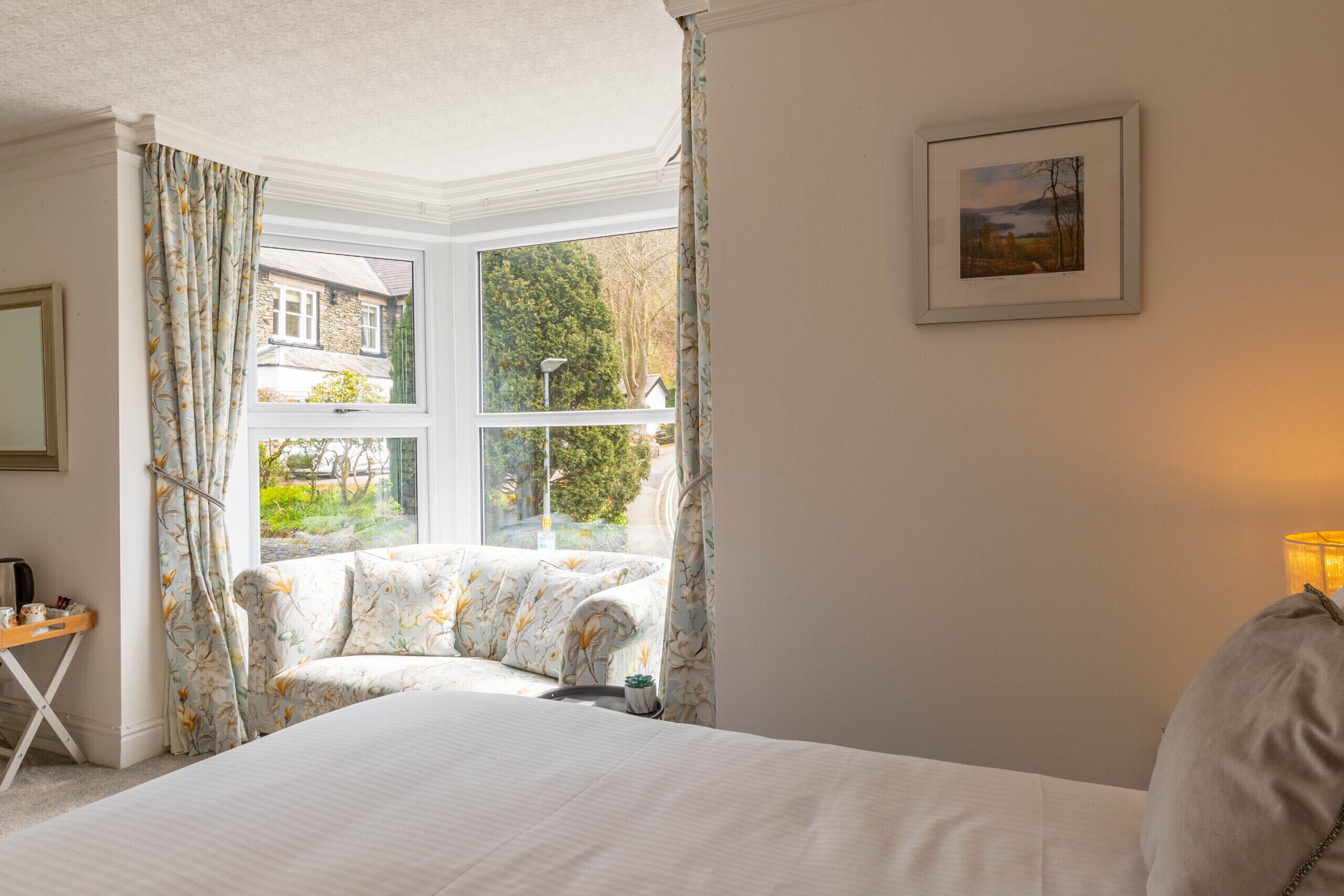 A bedroom with a large bed, a window with curtains, a small table with a lamp, and a framed picture on the wall.
