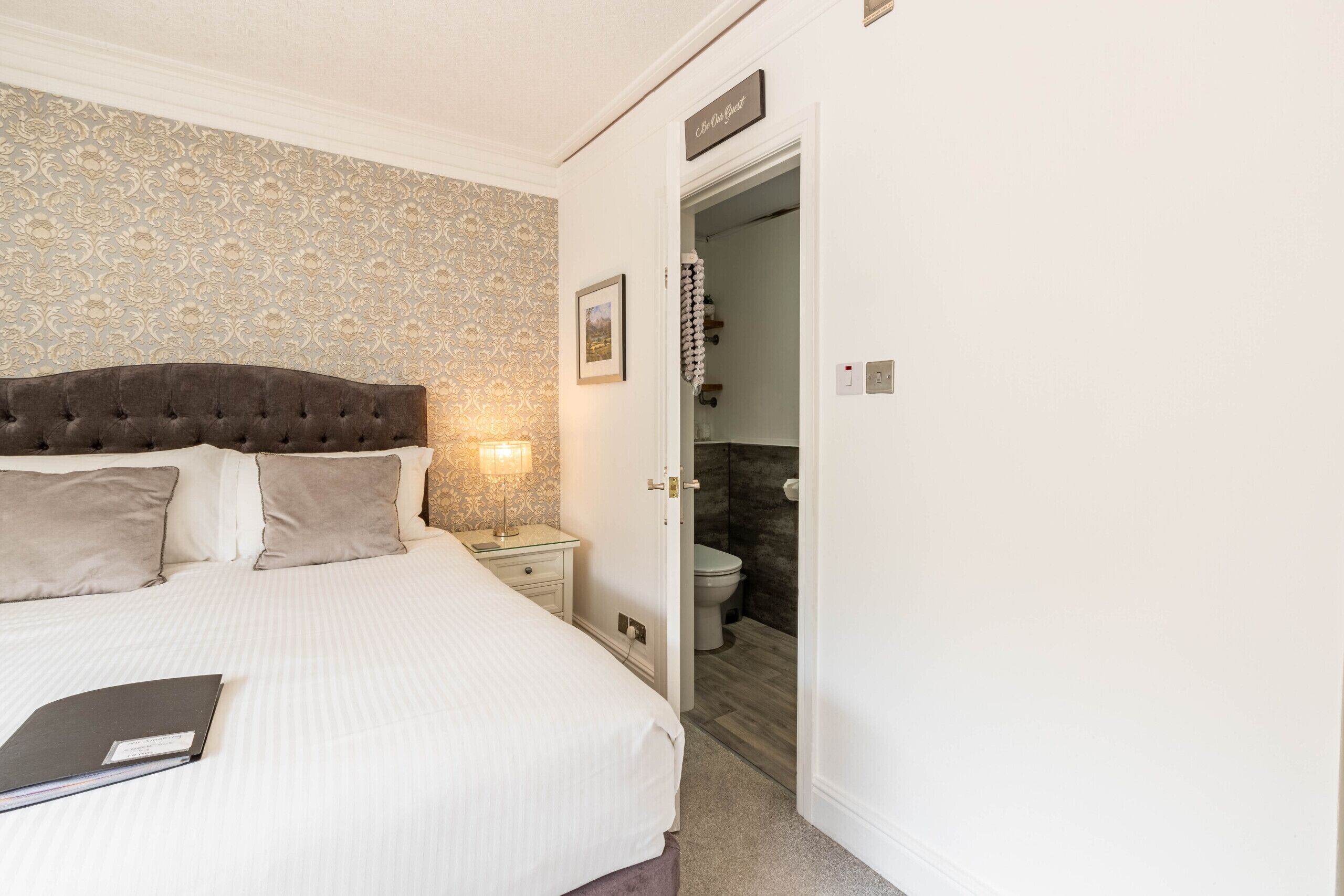 A bedroom with a large bed, a nightstand, a bedside lamp, and a door leading to a bathroom.