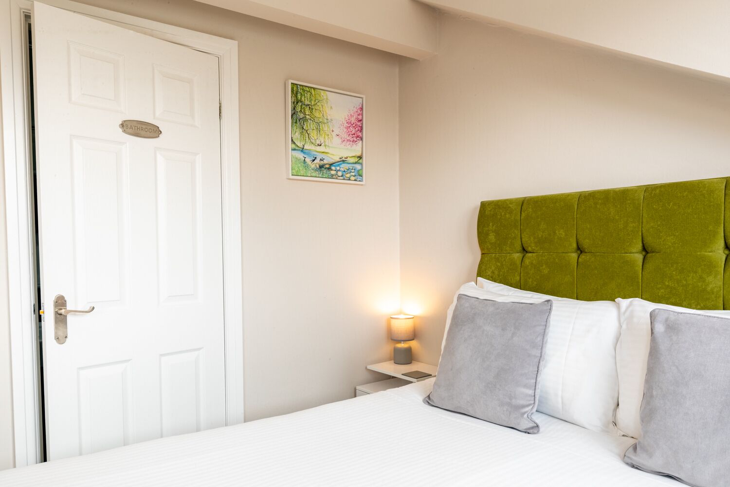 Une chambre avec une porte blanche, une tête de lit recouverte d’un tissu vert, une table de chevet avec une lampe et un tableau accroché au mur.