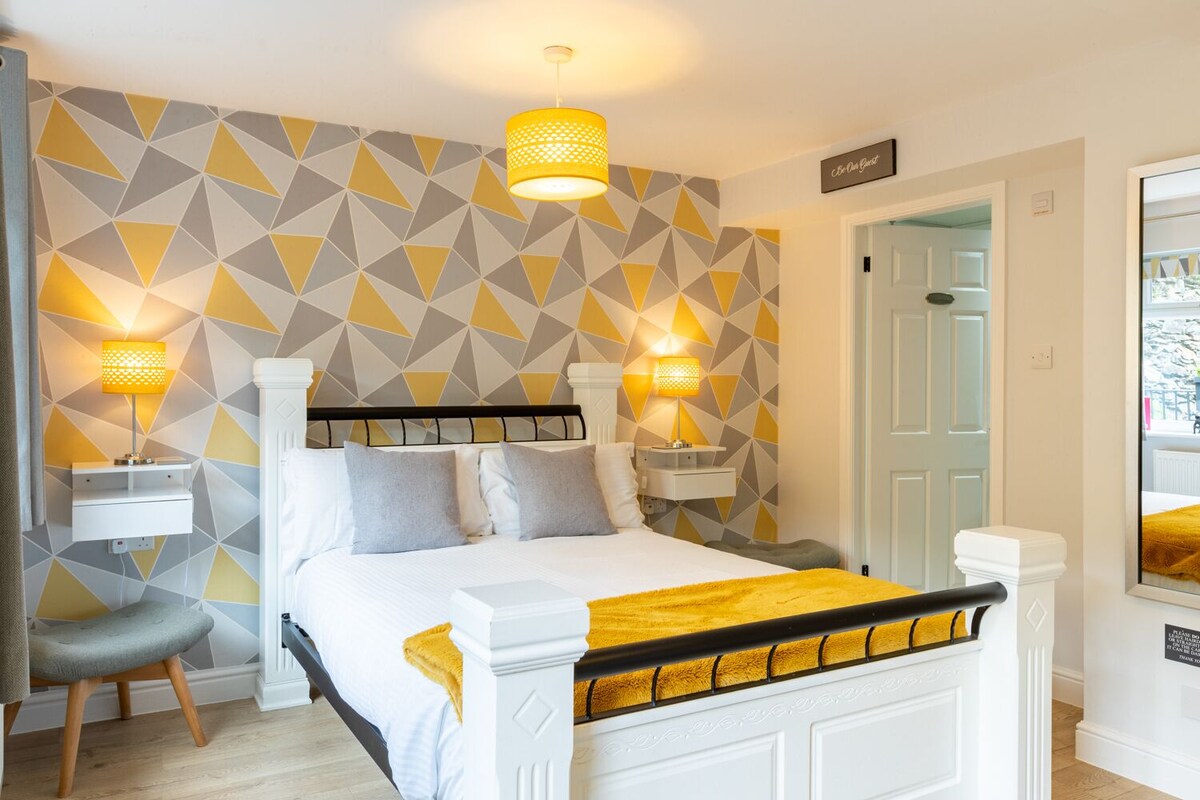 Un dormitorio con pared de estampado geométrico, una cama blanca con manta amarilla, lámparas de noche y una pequeña silla.