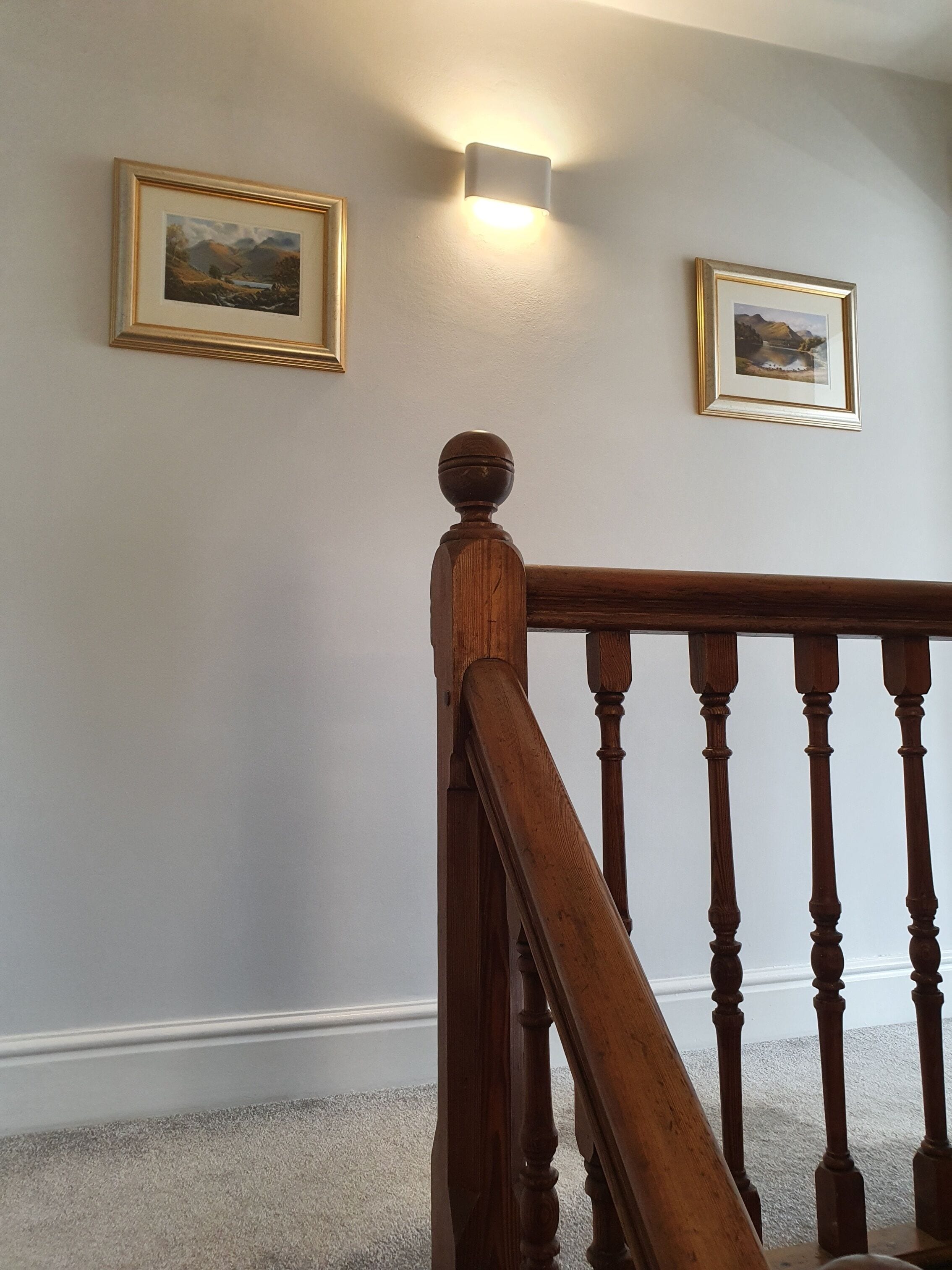 A staircase with wooden balusters and a handrail, leading to a landing with two framed landscape paintings on the wall.