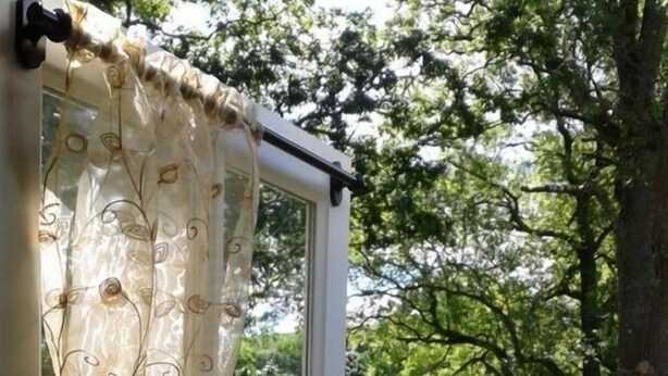 Ett fönster med en dekorativ gardin, utsikt över ett grönskande landskap och en eldstad utomhus.