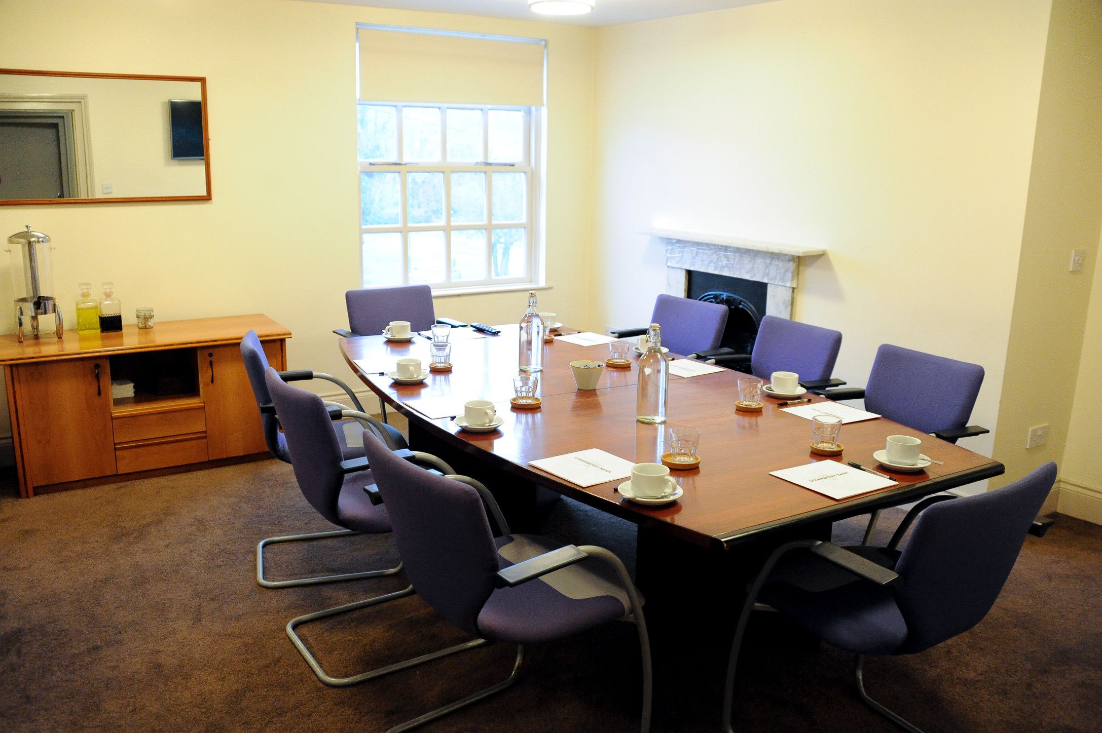 A conference room with a long table, chairs, a desk with a lamp, and a window.