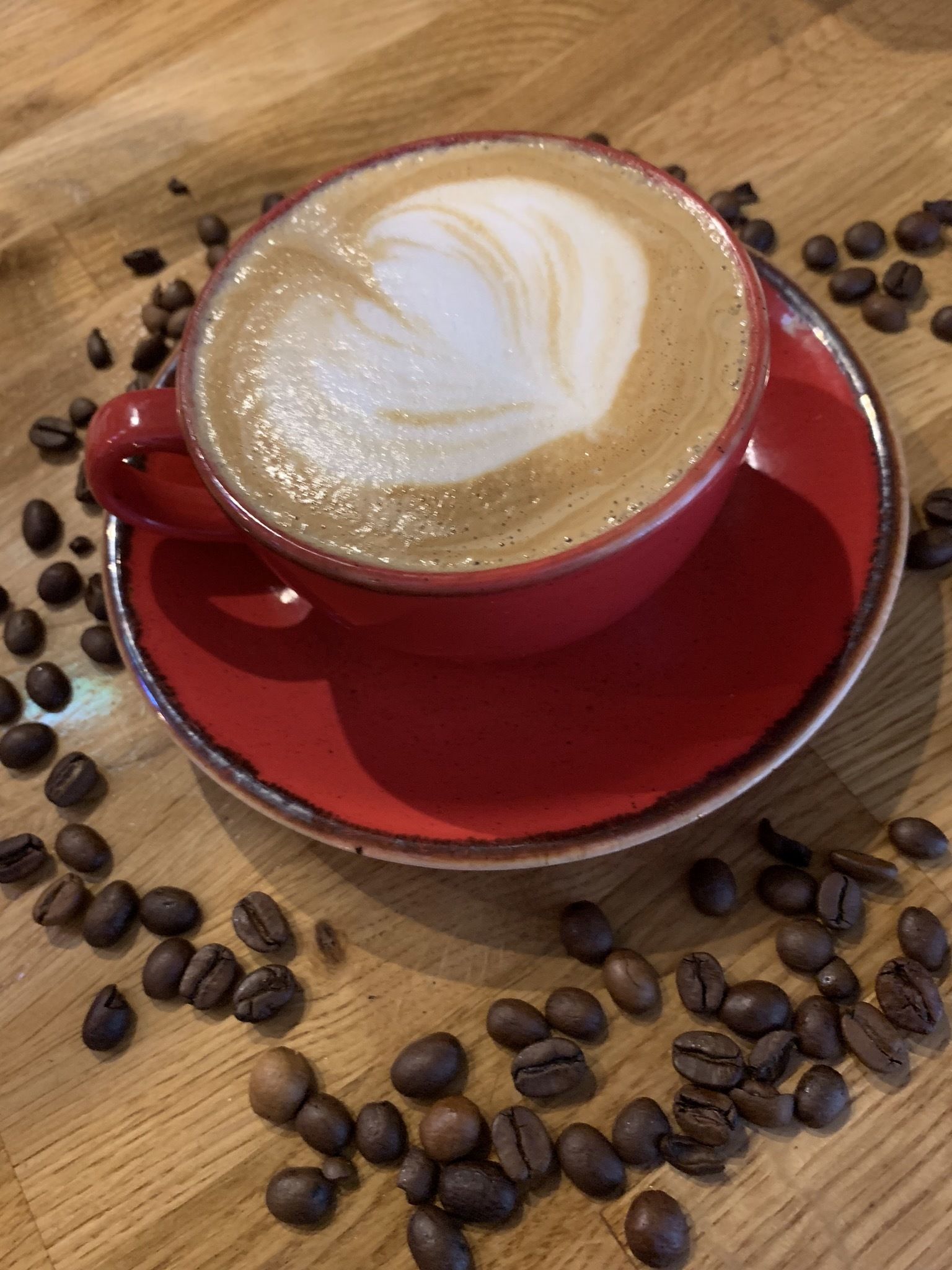 A red cup of coffee with latte art on a wooden table surrounded by coffee beans.