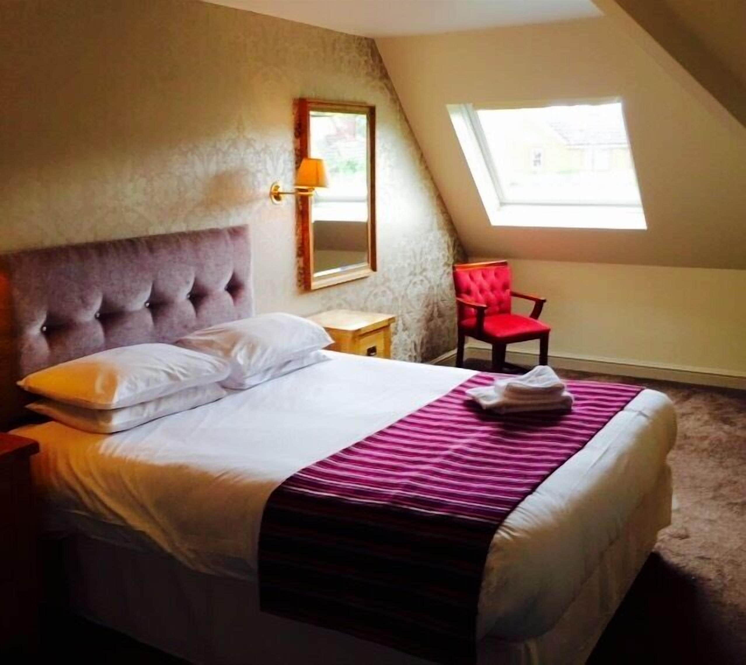 Una cama bien hecha con sábanas blancas y una manta rayada de color rosa, un sillón tapizado en rojo, una mesita de noche con una lámpara, y una ventana con vistas al exterior.