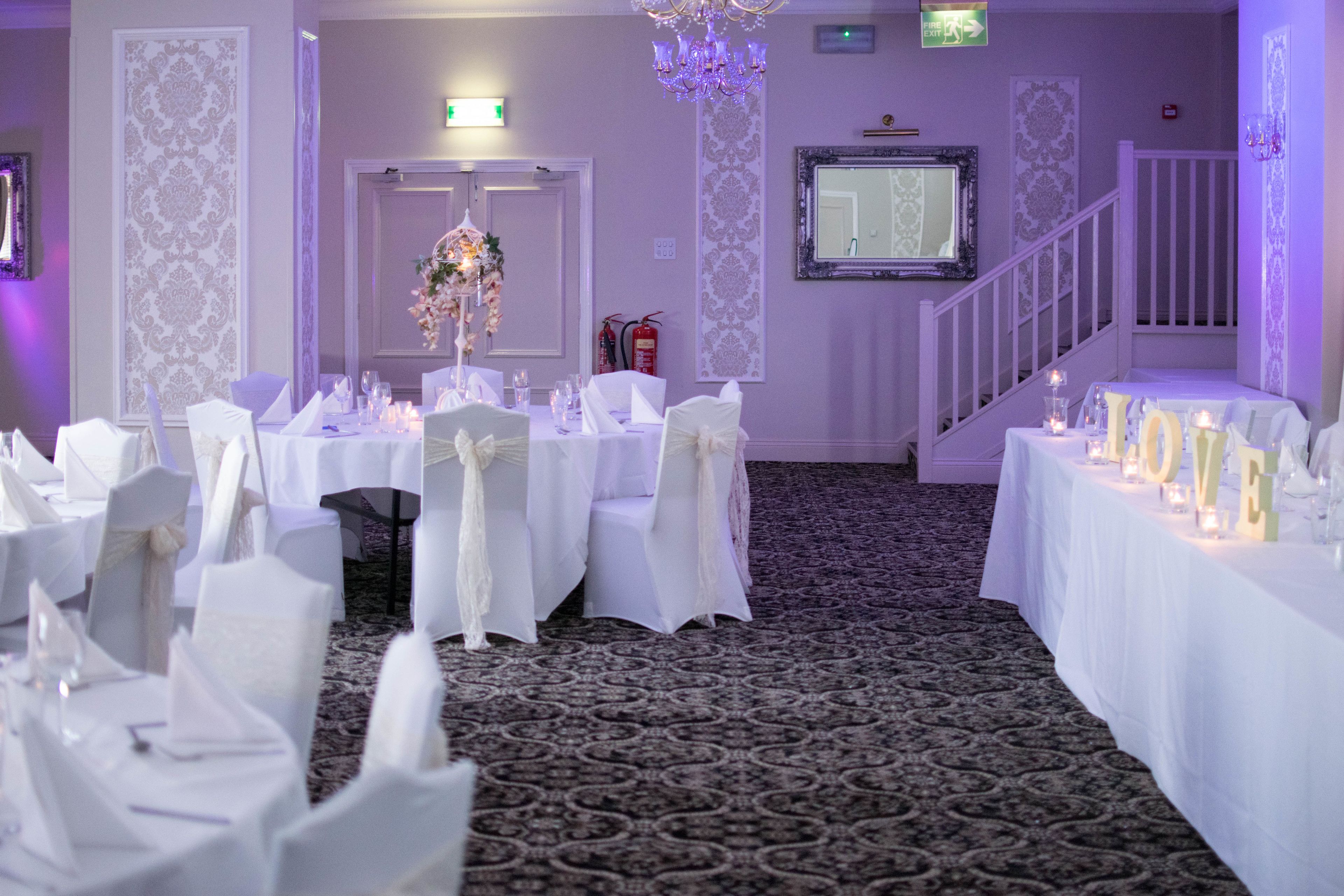 A banquet hall with tables set for a formal event, featuring white tablecloths, chairs with white sashes, and a chandelier.