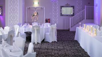 A banquet hall with tables set for a formal event, featuring white tablecloths, chairs with white sashes, and a chandelier.