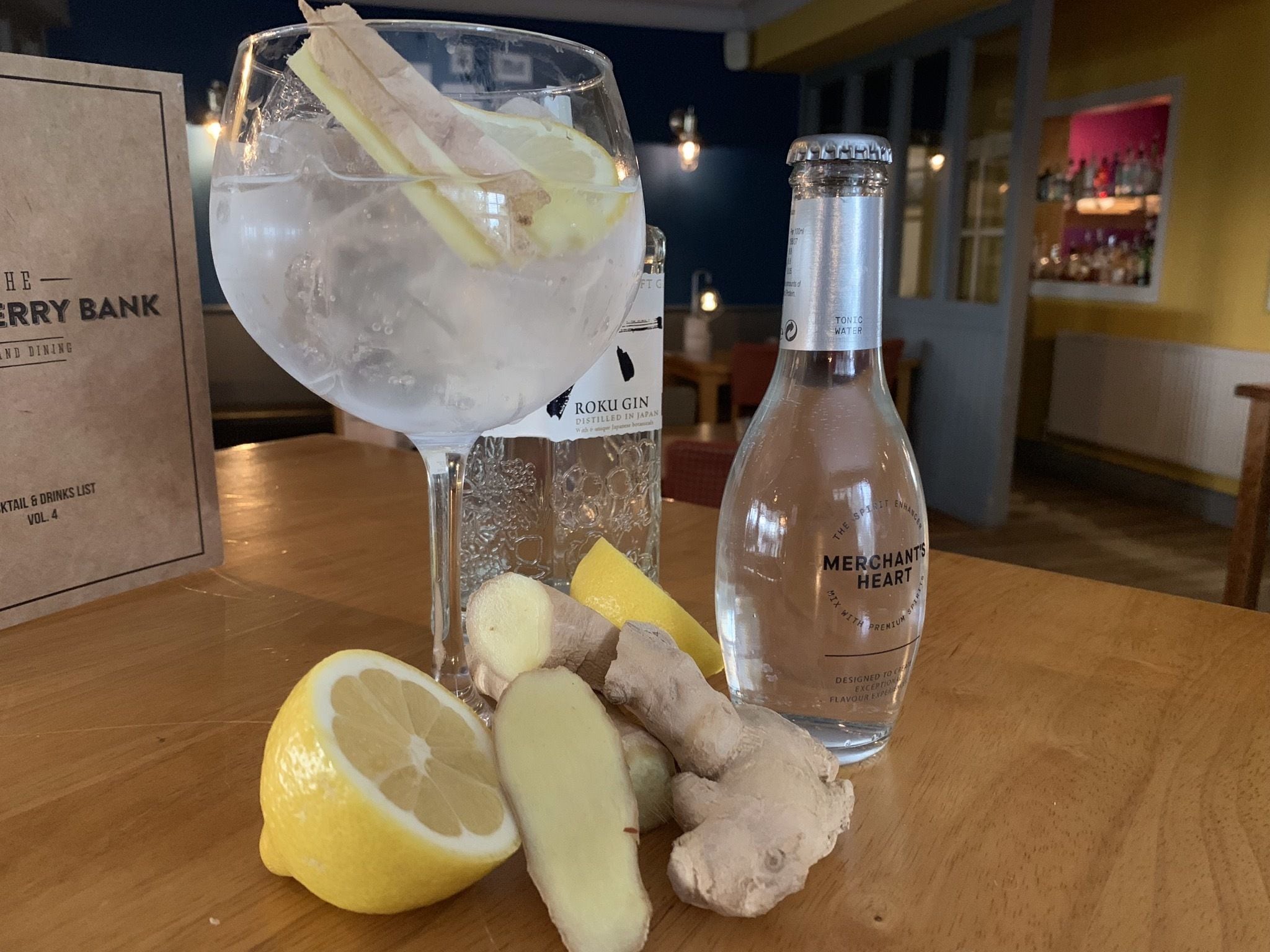 A cocktail glass with ice, lemon slices, and a ginger slice on a wooden table. A bottle labeled "Merchants Heart" is also visible.