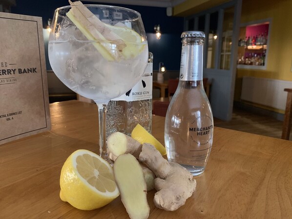 A cocktail glass with ice, lemon slices, and a ginger slice on a wooden table. A bottle labeled "Merchants Heart" is also visible.