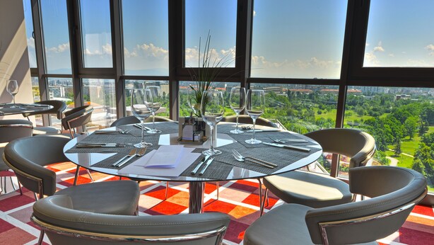 A round glass table with four chairs, a vase with a plant, and a city view through large windows. - Suite Hotel Sofia (Sofia)