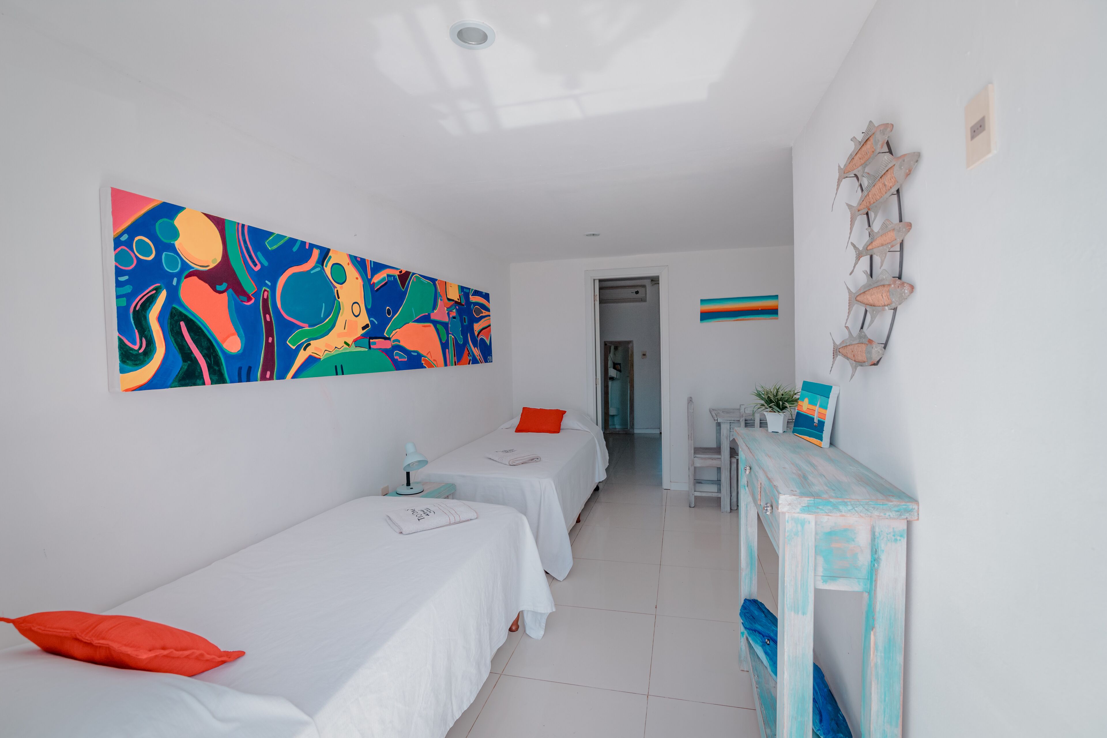 A room with two single beds, a desk, a colorful abstract painting, and a wall-mounted fish decoration.