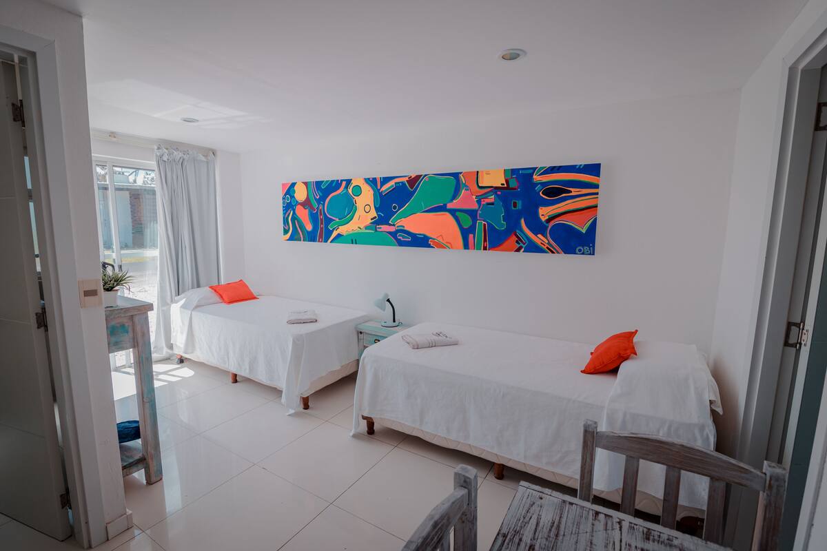 A hotel room with two beds, white bedding, a colorful abstract painting, and a view of the outdoors.