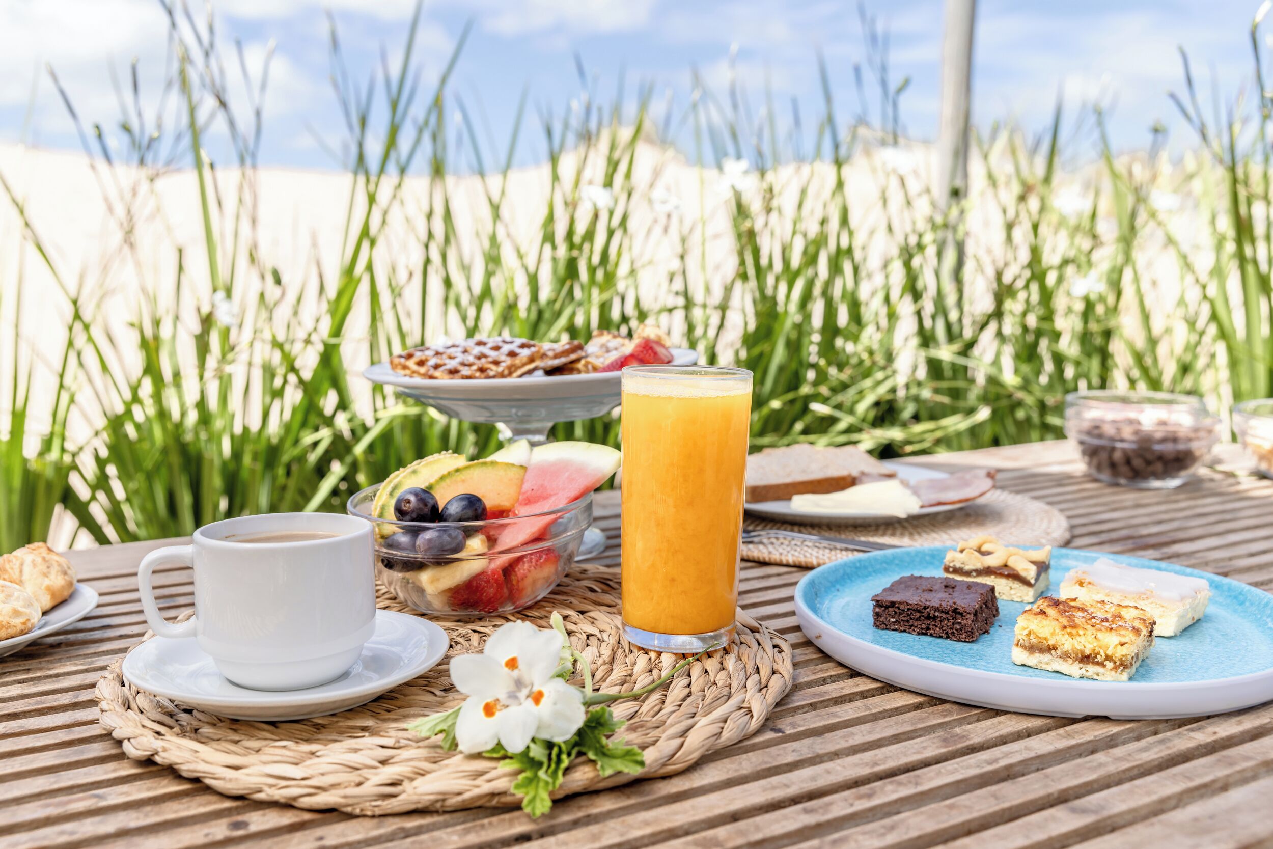 Desayuno con una taza de café, un vaso de jugo de naranja y diversas pastas sobre un tapete tejido al aire libre.