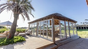 Un área de estar al aire libre techada, con piso de madera, paredes de vidrio y techo de paja.