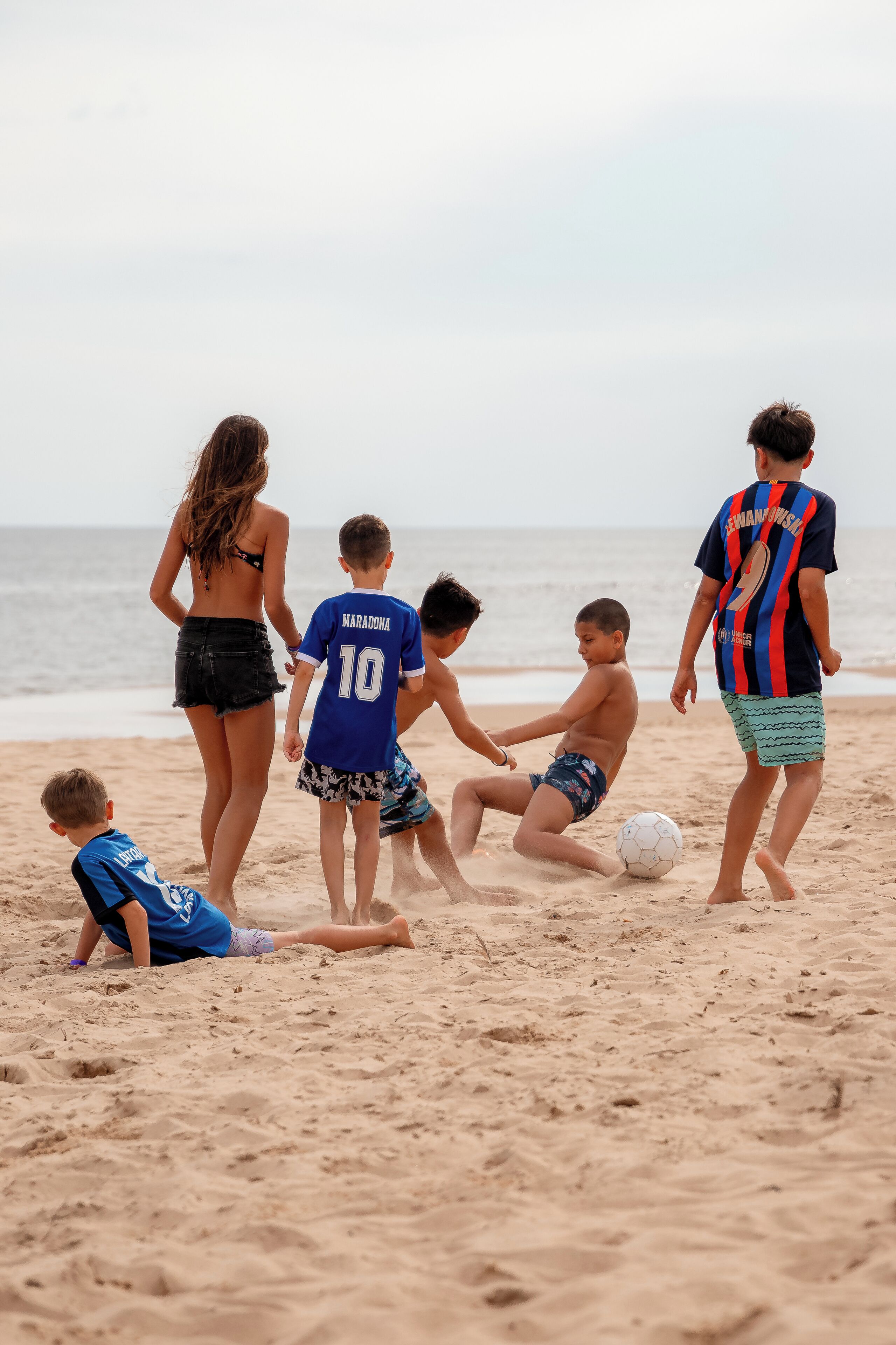A group of people playing soccer on a sandy beach with the ocean in the background.