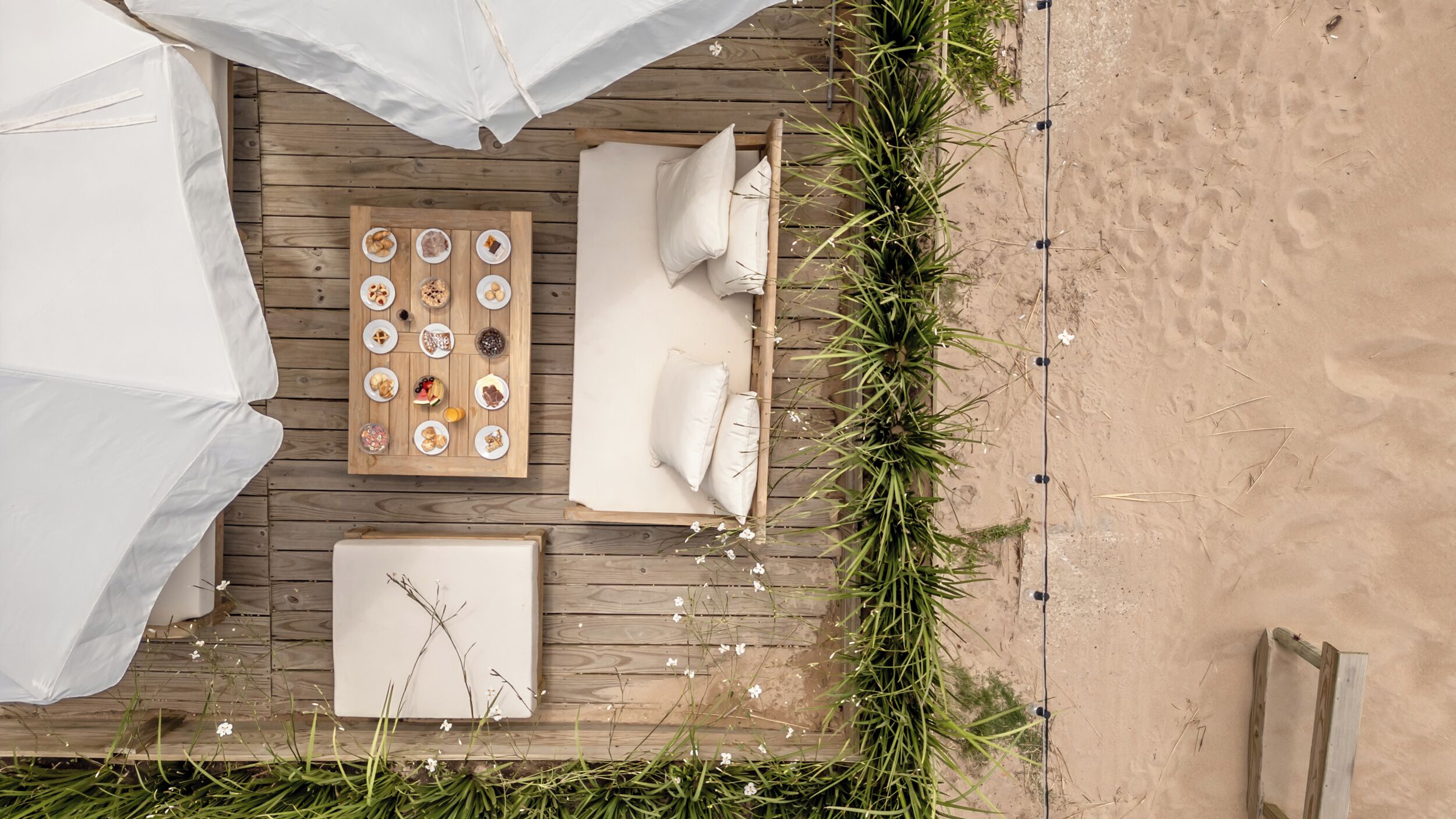 Un área de descanso frente a la playa con una terraza de madera, cojines blancos, una mesa con diferentes bocadillos y vistas a la playa.